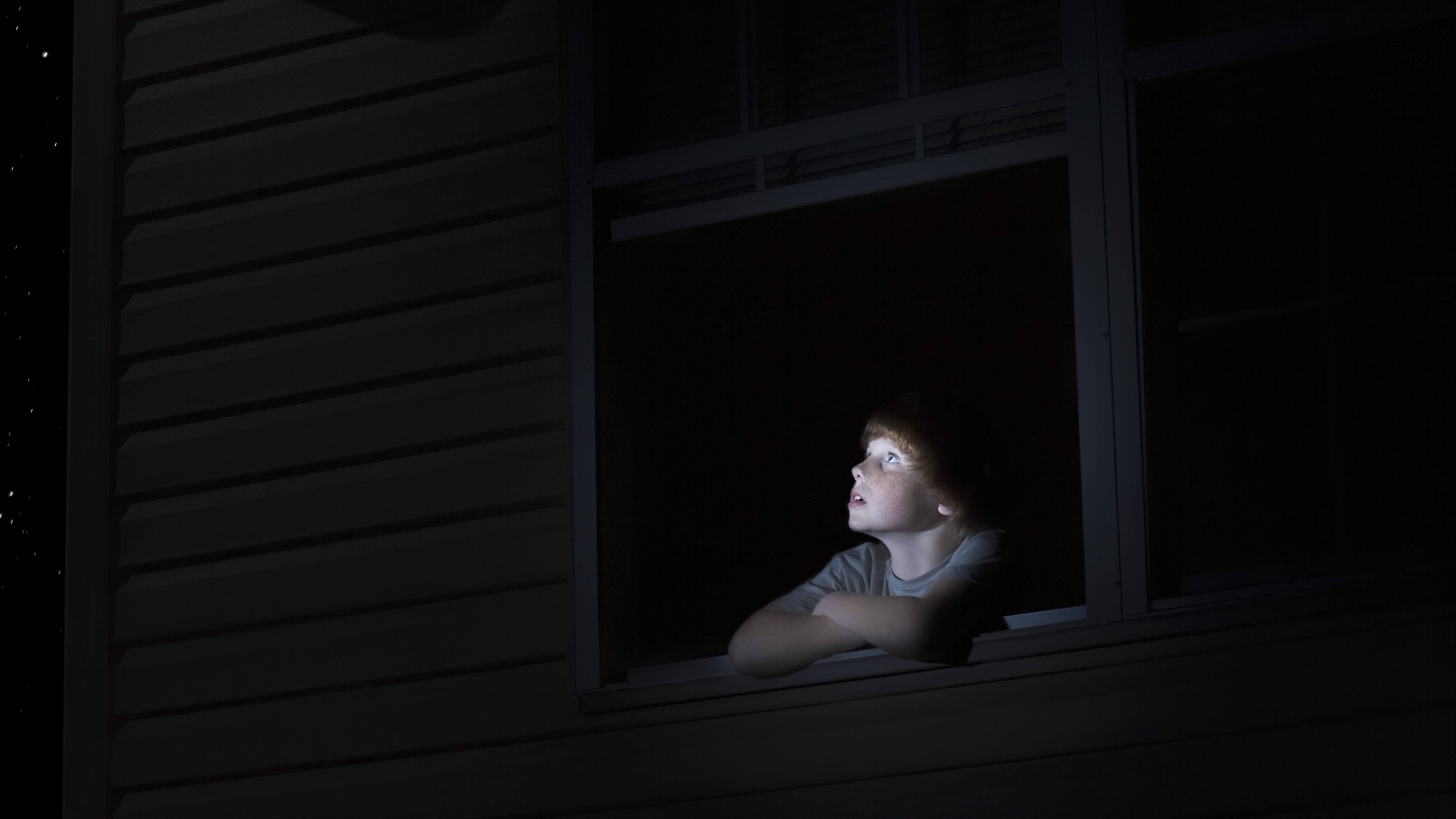 A boy gazes at the stars through a darkened window at night.